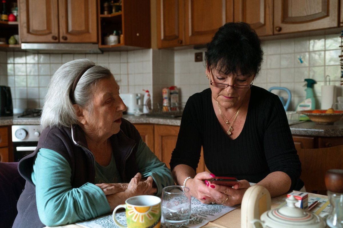 A caregiver sits beside an older woman at her kitchen table, helping her with a phone.