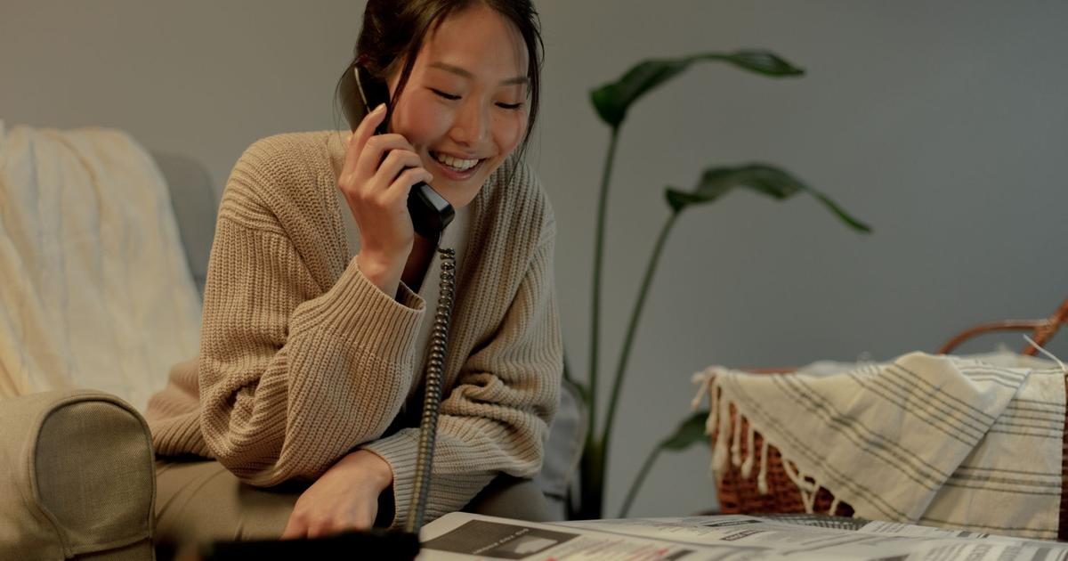A smiling young woman in a knit cardigan talks on a landline phone at home while a newspaper lies open on the table in front of her.