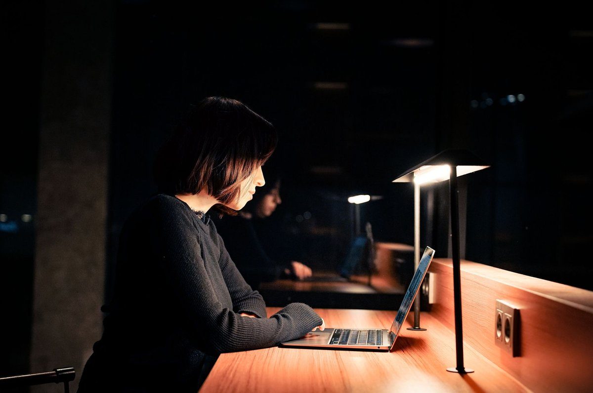 A woman works on a laptop at a desk lit by a small lamp late at night, when the office is closed but call-offs still come in.