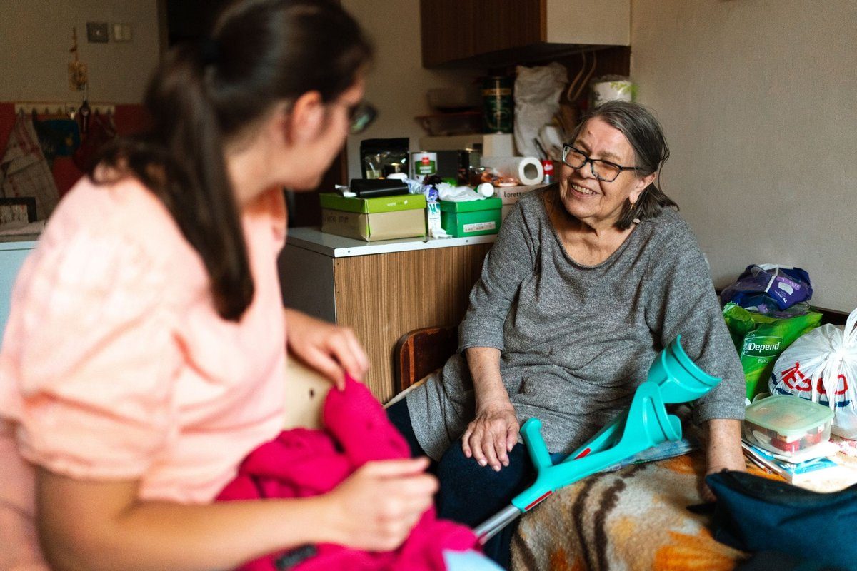 A caregiver in a pink top sits and talks with a smiling older woman who rests beside her crutch at home.