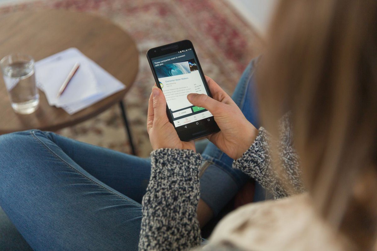 A woman in a knit cardigan sits in her living room reading a job listing on her smartphone.