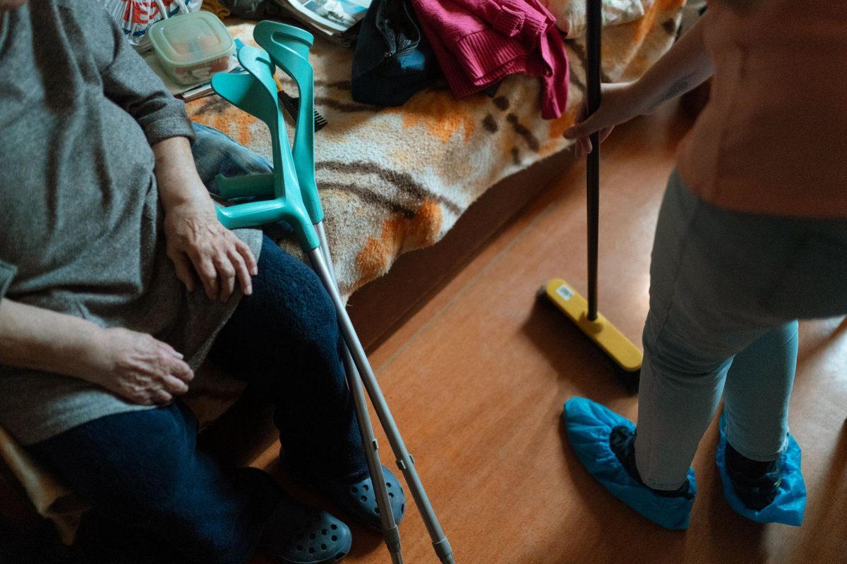 A caregiver in light blue shoe covers sweeps the floor next to an older woman seated with her forearm crutches.