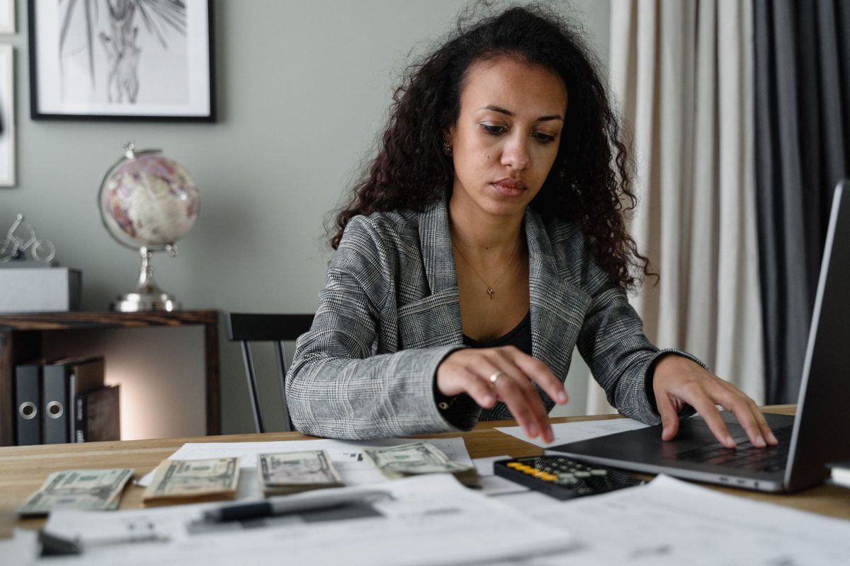 A woman in a gray plaid blazer works on a laptop at a desk covered with cash, receipts, and a calculator.