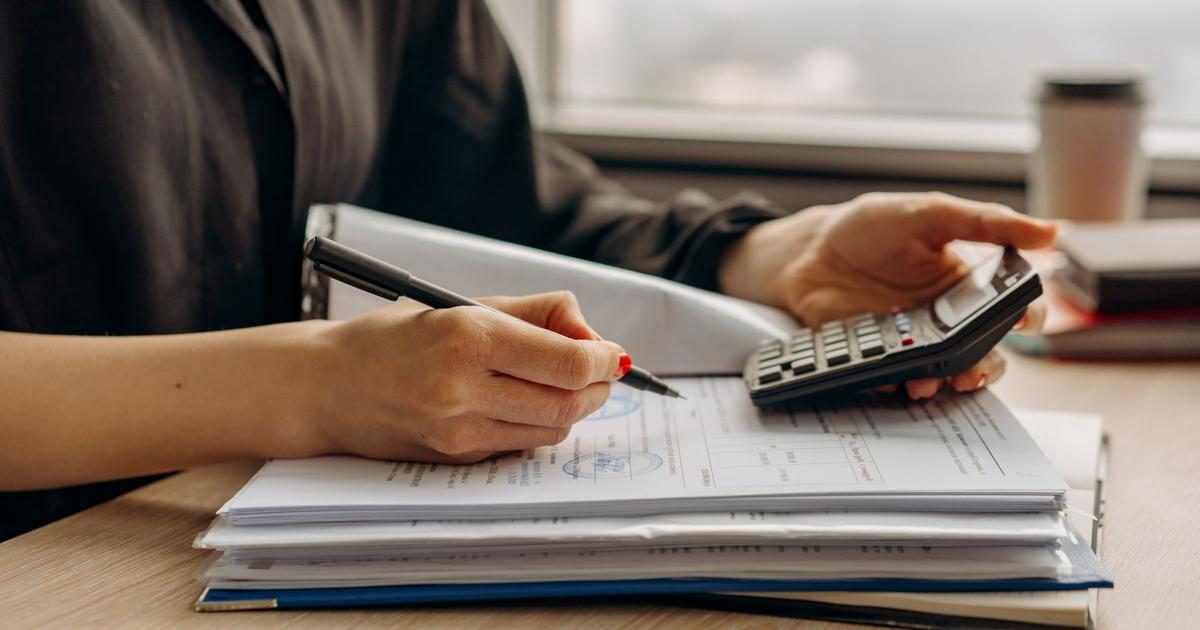 A person works through a stack of financial paperwork at a desk, holding a pen in one hand and a calculator in the other.