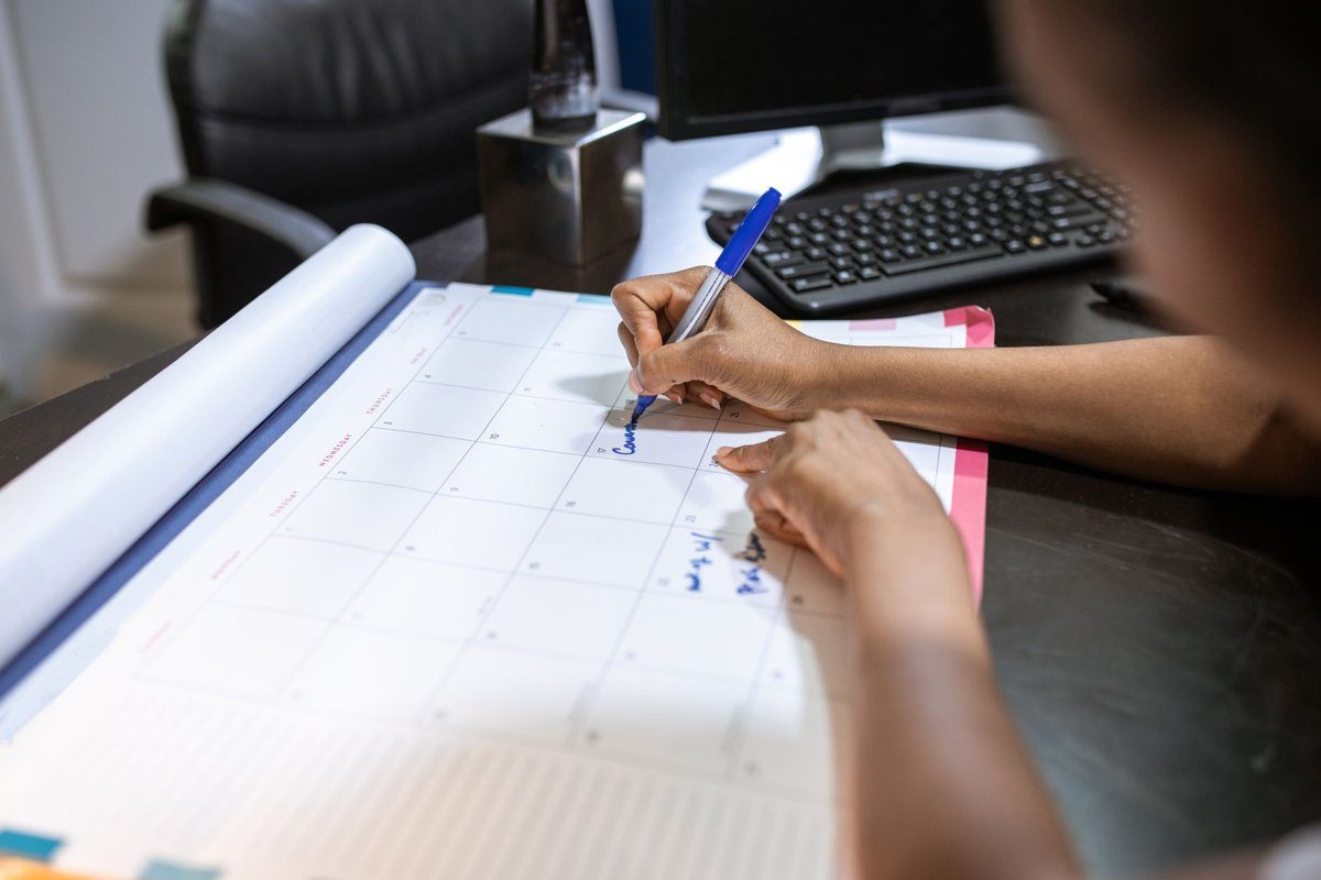 A scheduler writes on a large paper desk calendar with a blue pen next to a computer keyboard.