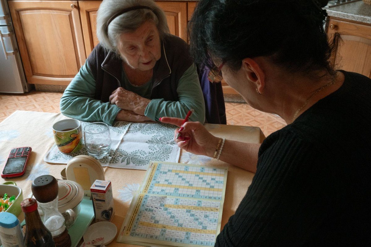 A caregiver and an older woman work on a crossword puzzle together at a kitchen table in the client's home.