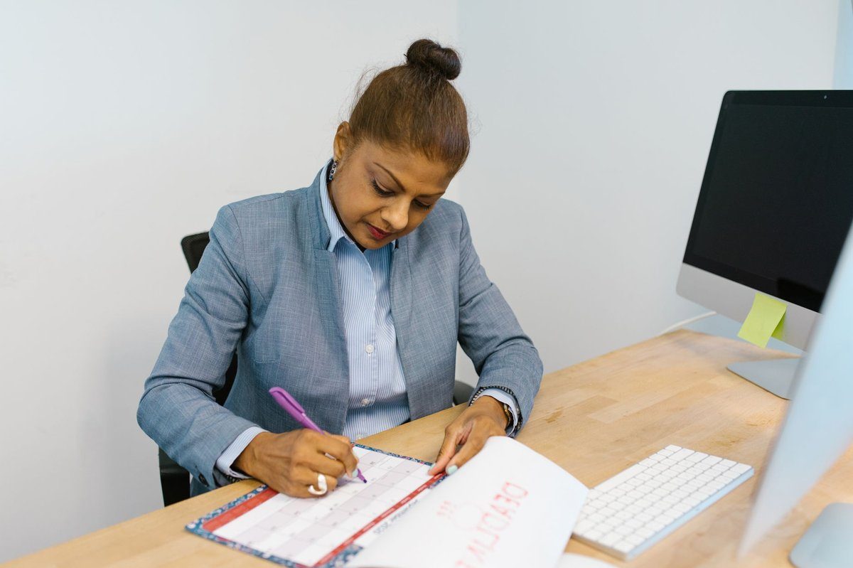An agency administrator in a gray blazer reviews paper records at her office desk beside a computer monitor.