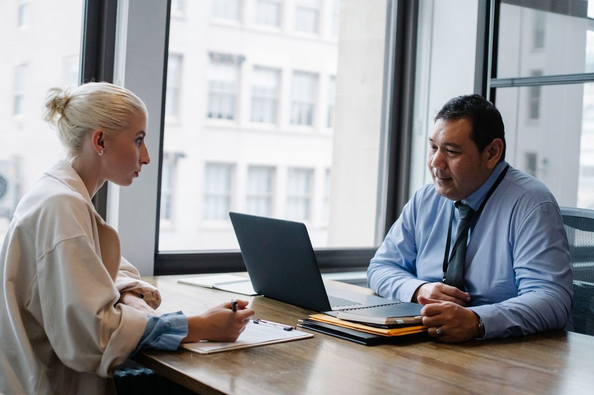 A job candidate fills in a form on a clipboard while a hiring manager with a lanyard listens across an office table.
