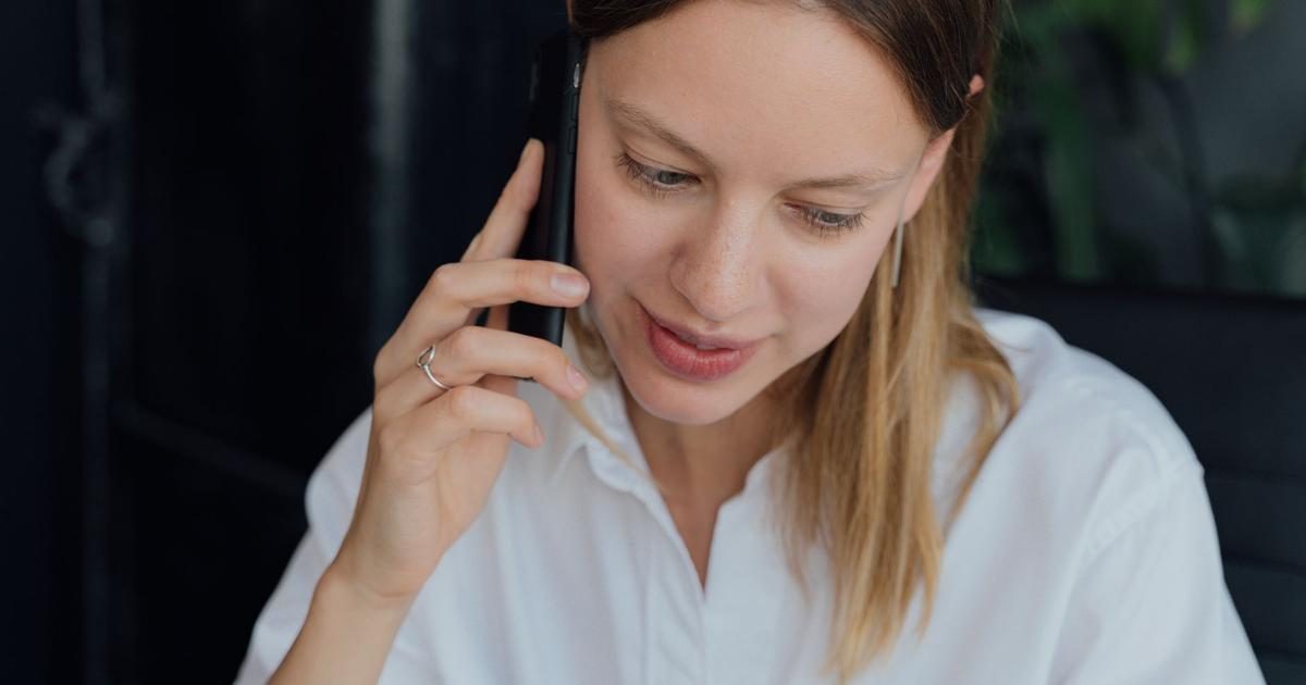 A young woman in a white shirt holds a smartphone to her ear and smiles slightly as she listens to a call.